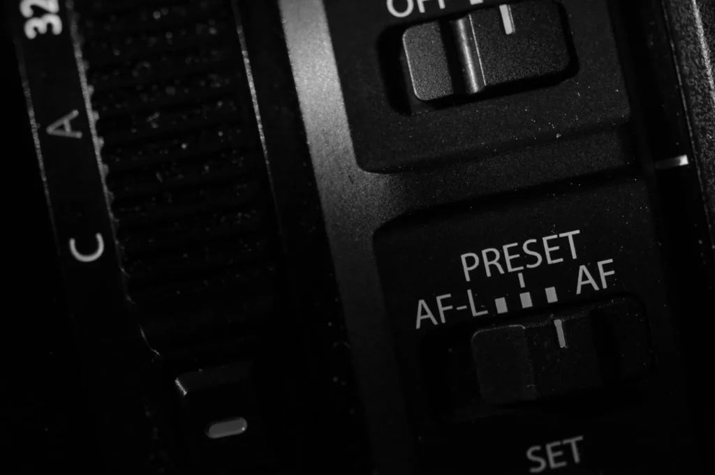 “Close-up of black camera lens controls with visible switches labeled "OFF," "PRESET," "AF-L," and "AF." The image conveys a technical, detailed mood.”