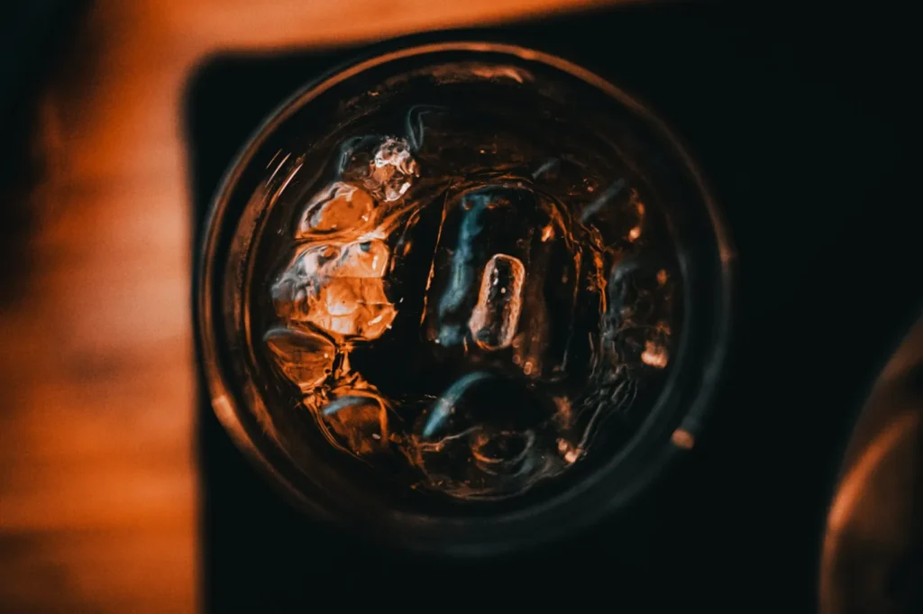 “Close-up of a dark beverage with ice in a glass, shot from above. The dim, warm lighting creates a moody and inviting atmosphere.”