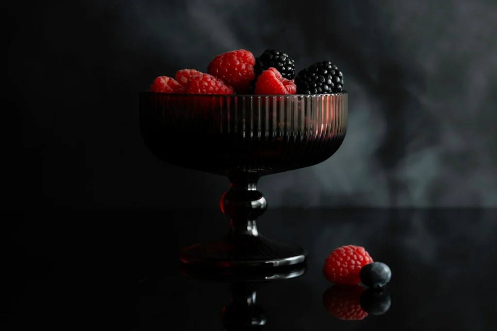 “A dark glass bowl filled with raspberries and blackberries sits on a reflective black surface. Smoky background and a few berries placed beside the bowl.”