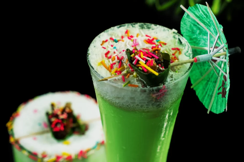 A tall glass of green shake topped with colorful sprinkles and a green umbrella. A small leaf and chocolate accent add a festive touch.