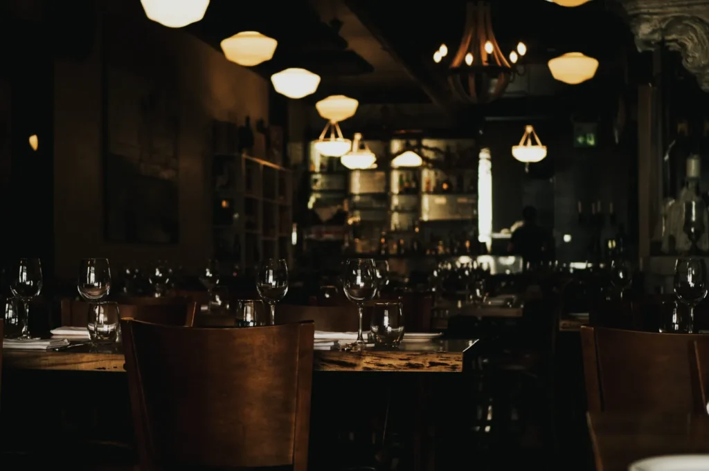Dimly lit restaurant interior with wooden tables set with wine glasses and napkins. A warm, inviting ambience with chandeliers and a visible bar in the background.