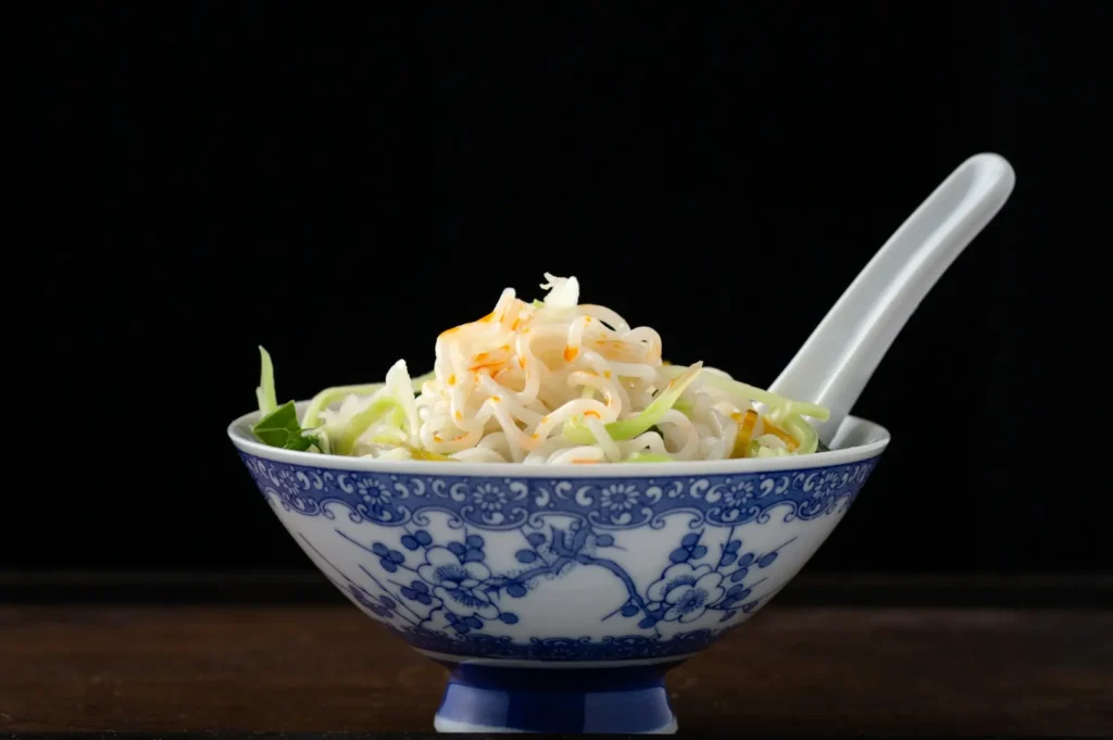 A blue and white porcelain bowl filled with noodles and green vegetables, topped with a touch of sauce. A white spoon rests in the bowl, set against a dark background.