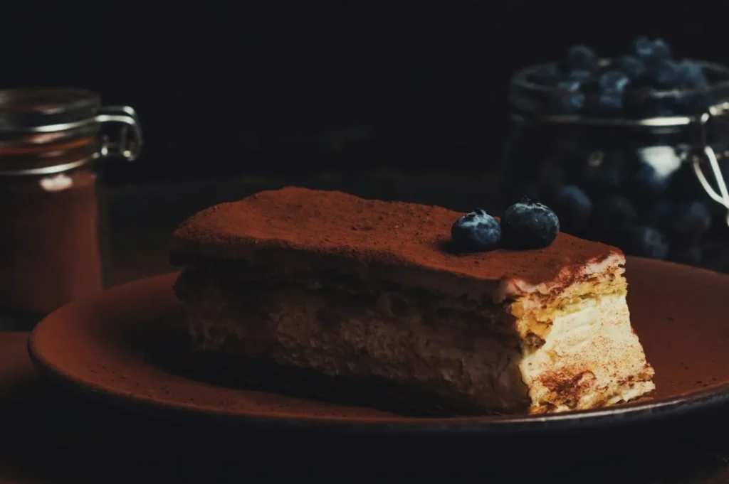 “A slice of creamy tiramisu dusted with cocoa powder is garnished with blueberries on a brown plate. A jar of blueberries and cocoa appear blurred in the background.