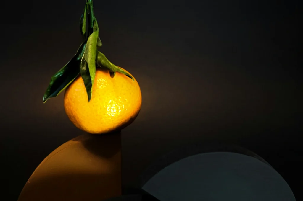 “A ripe orange mandarin with green leaves sits atop a geometric shape against a dark background. The composition is minimalistic and elegant.”