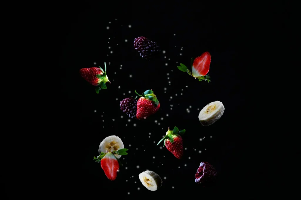 “Assorted fruits, including strawberries, blackberries, raspberries, and banana slices, appear to float on a dark background, creating a fresh and vibrant scene.”