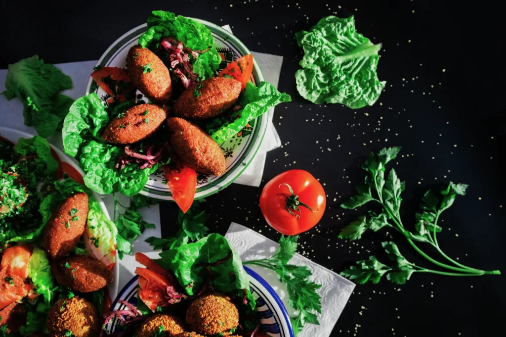 “Plates of falafel on leafy greens and sliced tomatoes, with fresh parsley and a whole tomato nearby. Bright colors and fresh ingredients on a dark background.”
