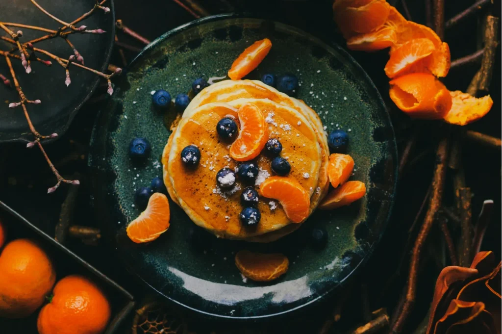 A stack of pancakes on a dark green plate, garnished with blueberries and tangerine slices, surrounded by rustic twigs and whole tangerines.