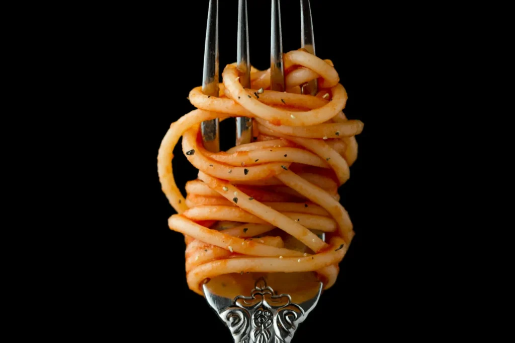 “A silver fork artfully holds coiled spaghetti with tomato sauce, sprinkled with black pepper, set against a dramatic black background.”