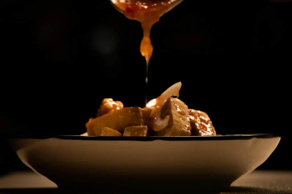 Sauce is drizzled over a dish of tofu and vegetables on a plate. The dark background highlights the golden hue of the sauce, creating a warm, appetizing mood.