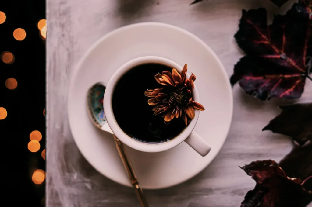“A cup of dark coffee sits on a saucer, adorned with a brown flower. A gold spoon rests inside. Purple leaves and warm bokeh lights create an autumn feel.”
