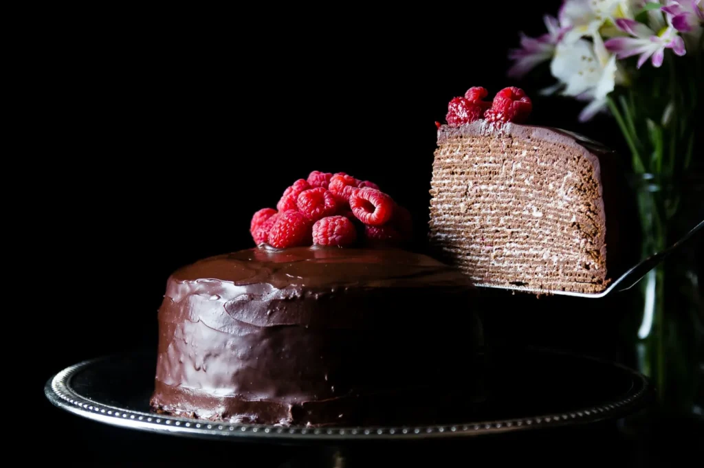 A chocolate cake with smooth ganache and raspberries on top is on a silver platter. A slice is cut to reveal multiple thin layers. Flowers are blurred in the background.