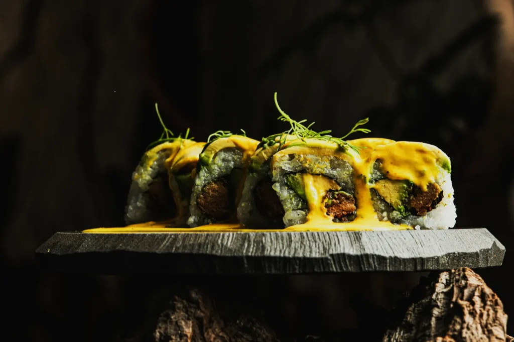 Close-up of sushi rolls topped with a creamy yellow sauce and garnished with sprigs of herbs. The sushi rests on a textured stone slate, set against a dark, moody background.