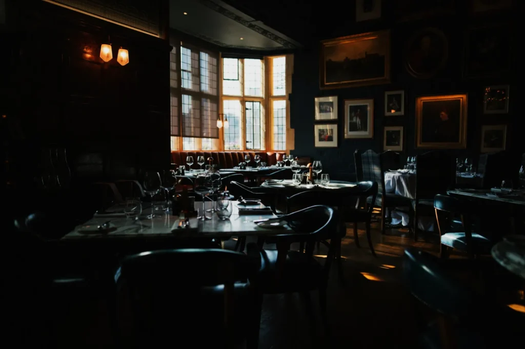 Dimly lit, elegant restaurant interior with dark wooden tones, large windows, and framed art. Tables are set for fine dining, creating a cozy, upscale atmosphere.