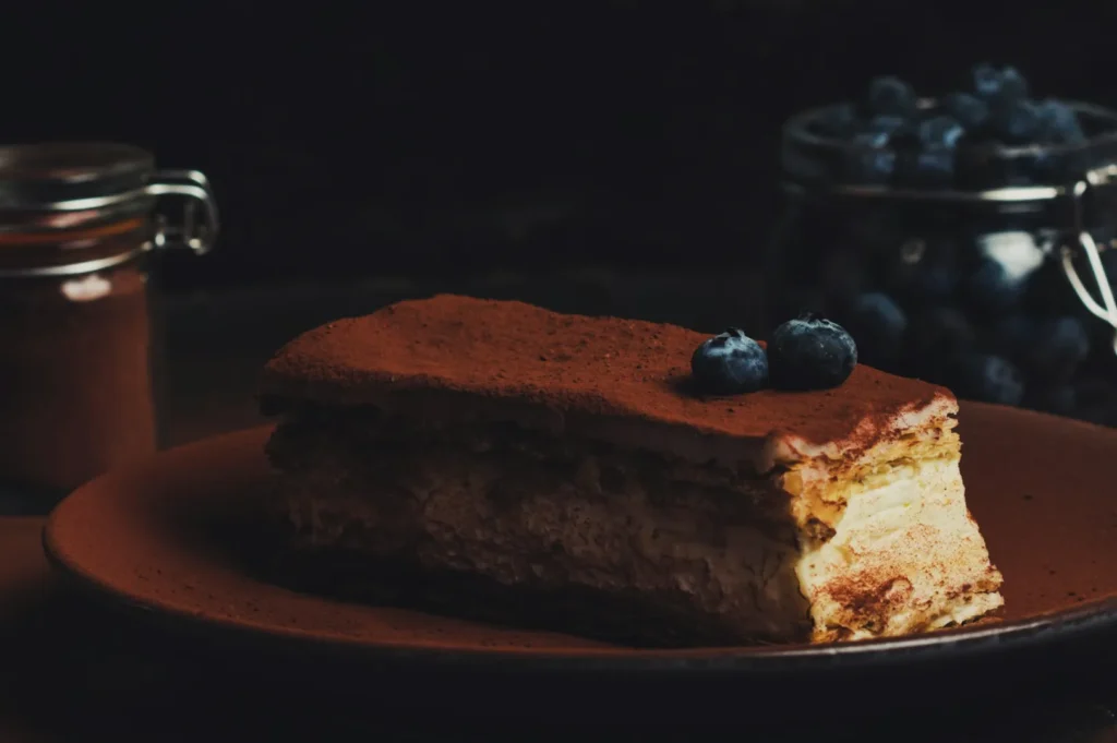 A slice of tiramisu topped with blueberries on a brown plate. In dim lighting, a jar of cocoa and a jar of blueberries are blurred in the background.