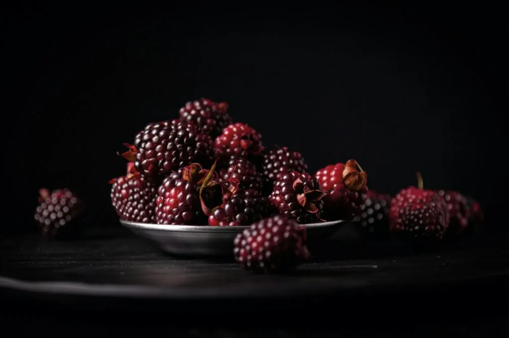“A plate of ripe blackberries on a dark surface, set against a black background. The berries glisten with a rich, deep purple hue, evoking natural freshness.”