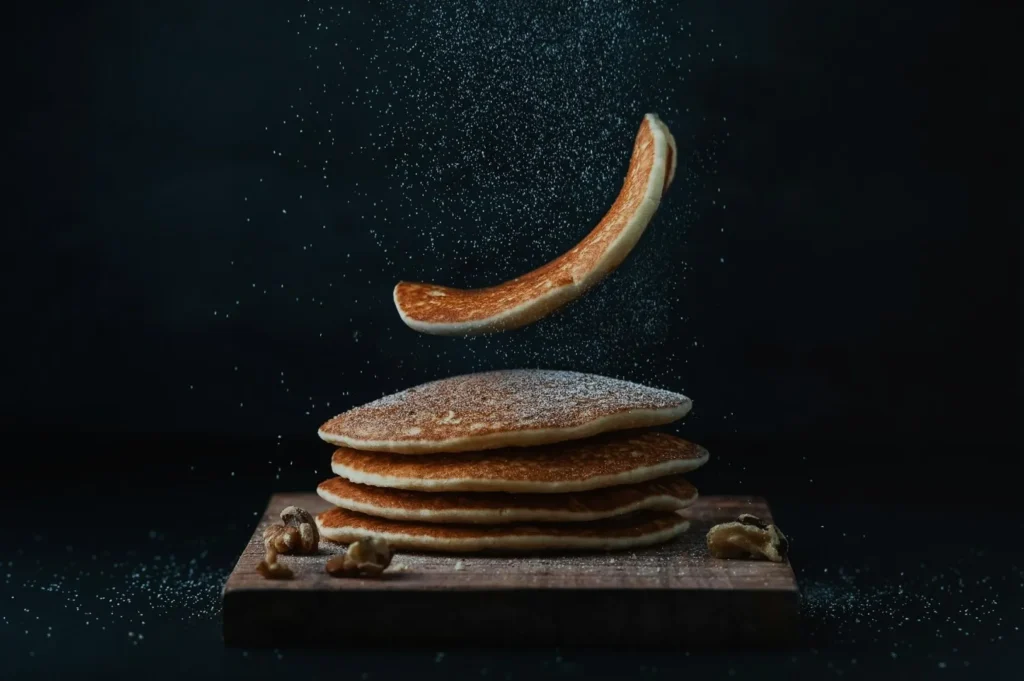 “A stack of pancakes on a wooden board with powdered sugar gently falling over them. One pancake is flipping mid-air, with walnuts beside the stack.”