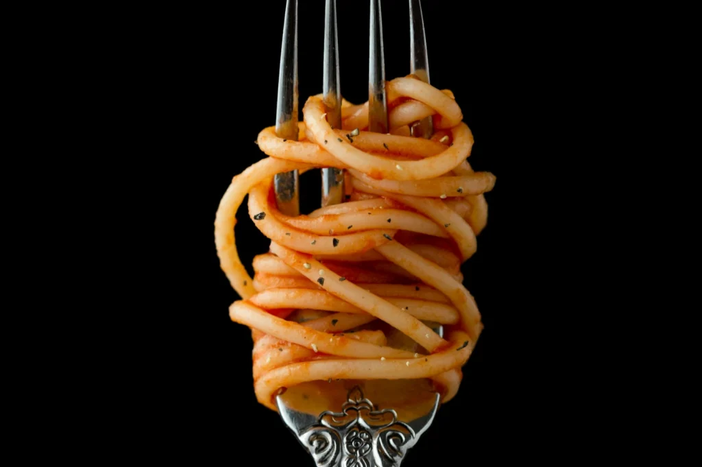 “A silver fork with intricately detailed handle holds twirled spaghetti covered in tomato sauce and pepper, set against a black background.”