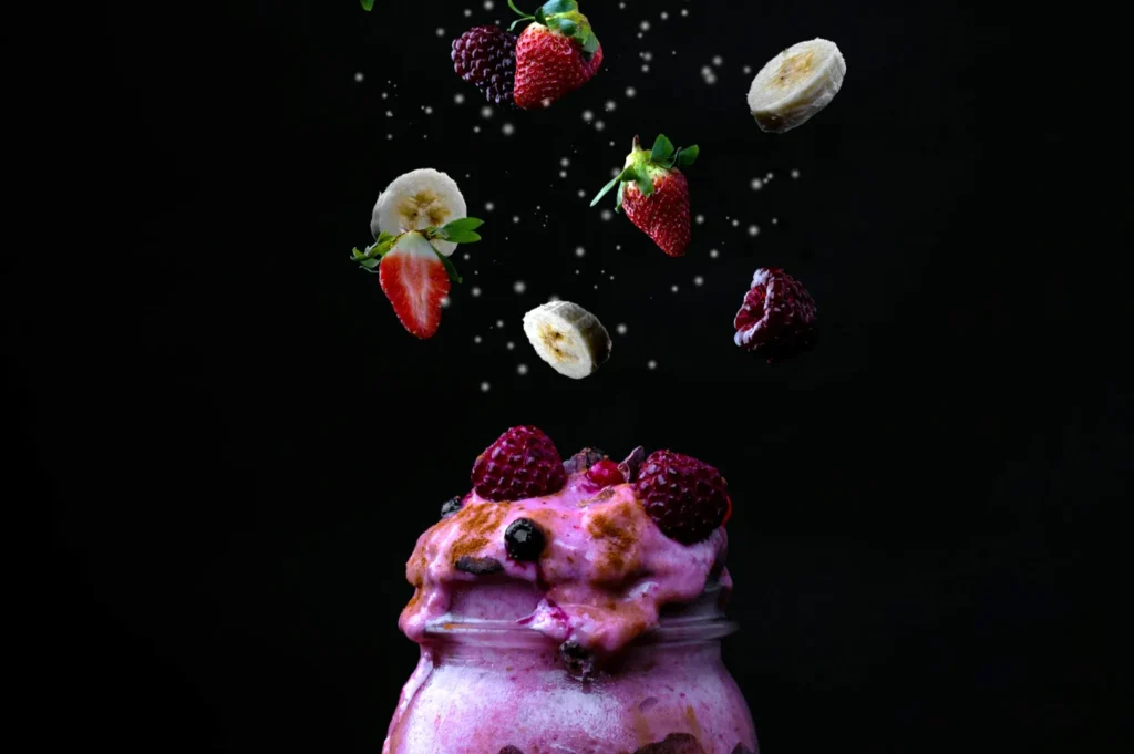 “A vibrant smoothie overflows a jar, topped with berries. Bananas, strawberries, and raspberries float above on a dark background, creating a lively scene.”