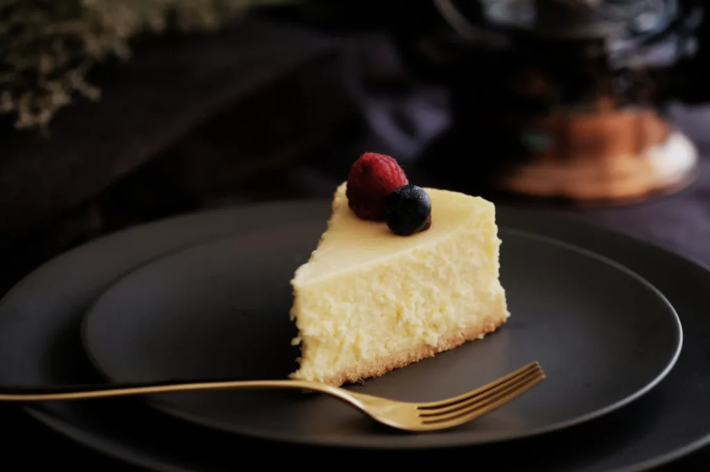 “A creamy cheesecake slice topped with a raspberry and blueberry sits on a dark plate, next to a gold fork. The setting feels elegant and inviting.”