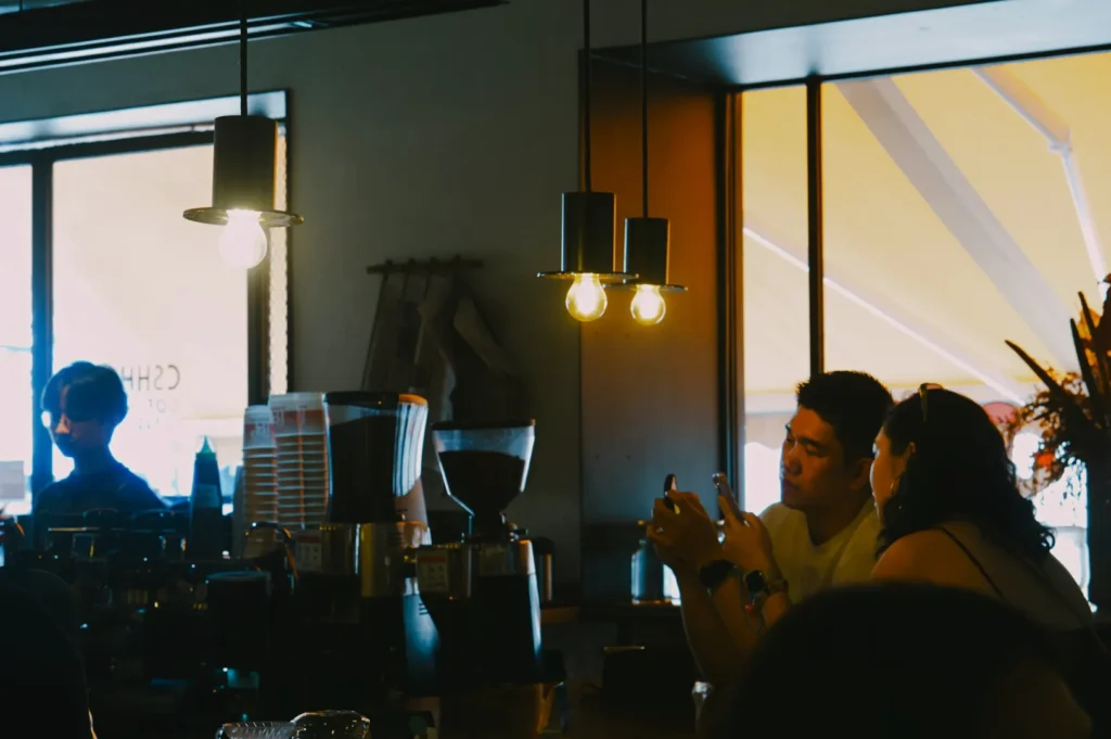 A cozy cafe scene with warm lighting shows two people seated at a counter lit by pendant lights, engaging with a smartphone. A barista works in the background.