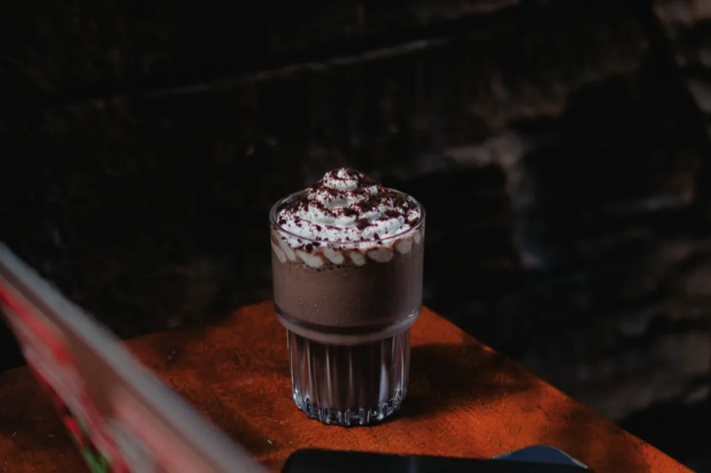 A creamy chocolate milkshake topped with whipped cream sits on a wooden table. The setting is cozy and dimly lit, evoking a warm, inviting atmosphere.