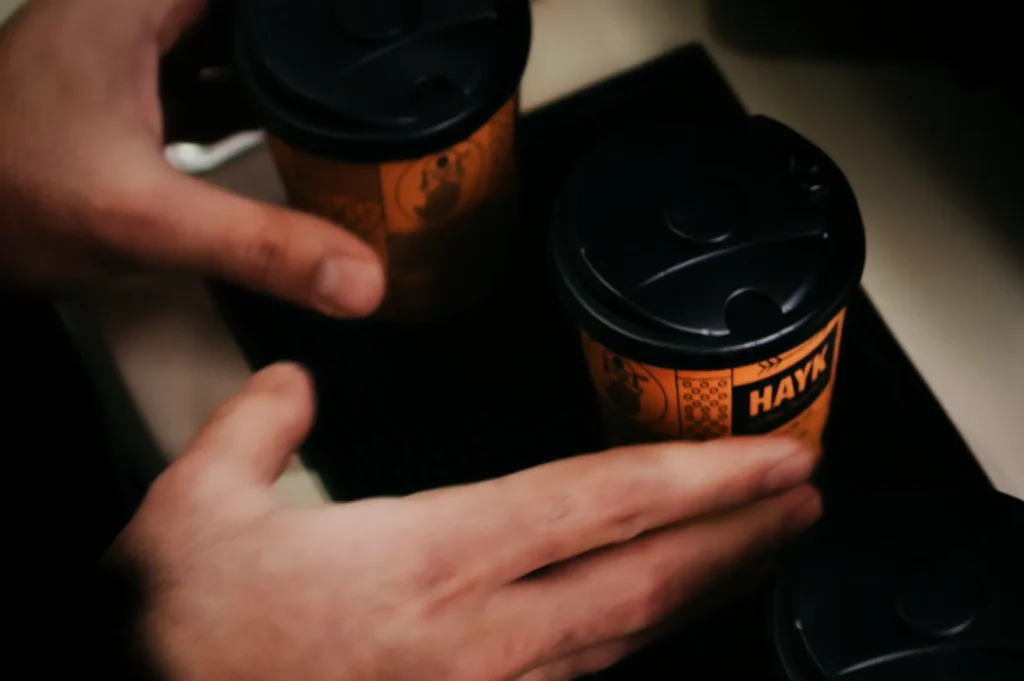 “Close-up of hands placing three orange coffee cups with black lids on a black tray. The cups have a playful logo and the word "HAYK".”