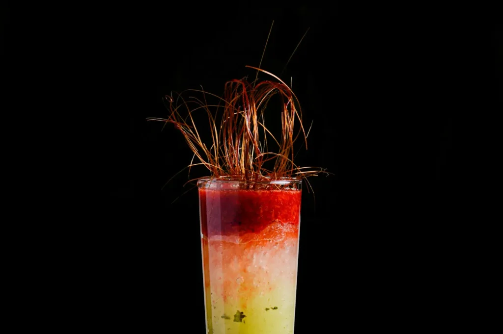 A vibrant cocktail in a glass against a black background. The drink transitions from yellow at the bottom to red at the top, topped with sprigs resembling copper wire, evoking a playful, artistic vibe.