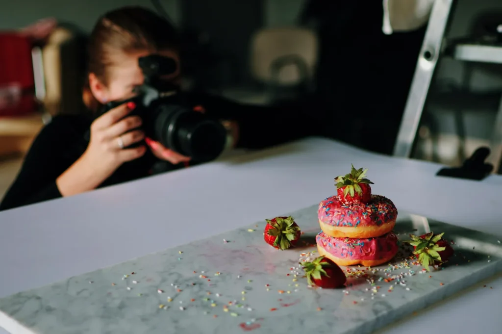 “A photographer focuses on a stack of two donuts topped with sprinkles and strawberries on a marble slab. The setup conveys a playful, creative vibe.”