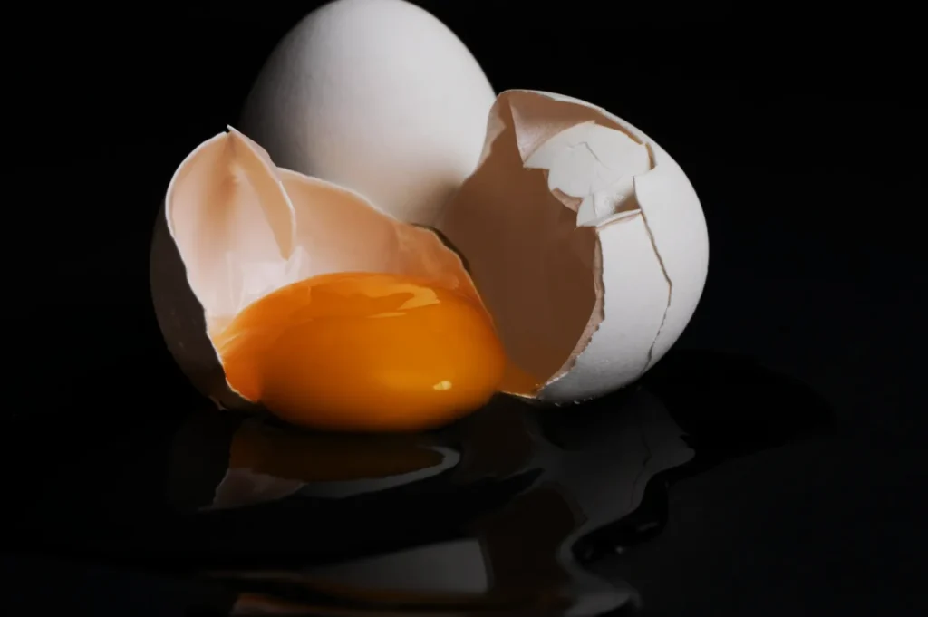 “Two eggs on a dark surface: one whole, one cracked open with a vibrant yellow yolk and clear egg white spilling out, creating a striking contrast.”