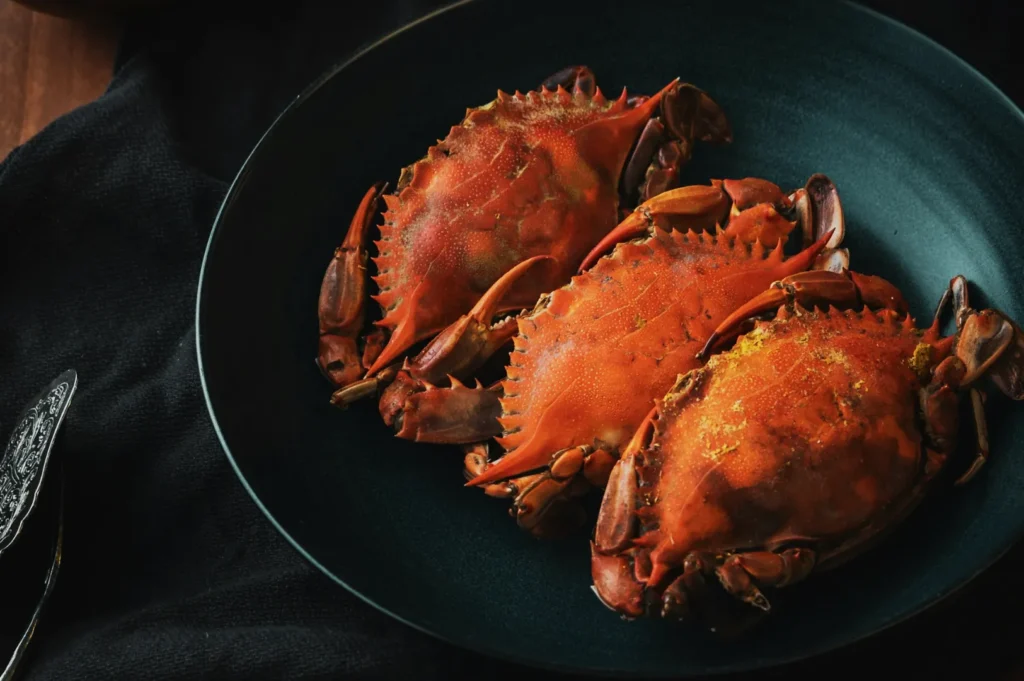 Three cooked crabs with rich, orange shells lie on a dark teal plate. The setting is on a textured black fabric, creating a rustic, elegant tone.