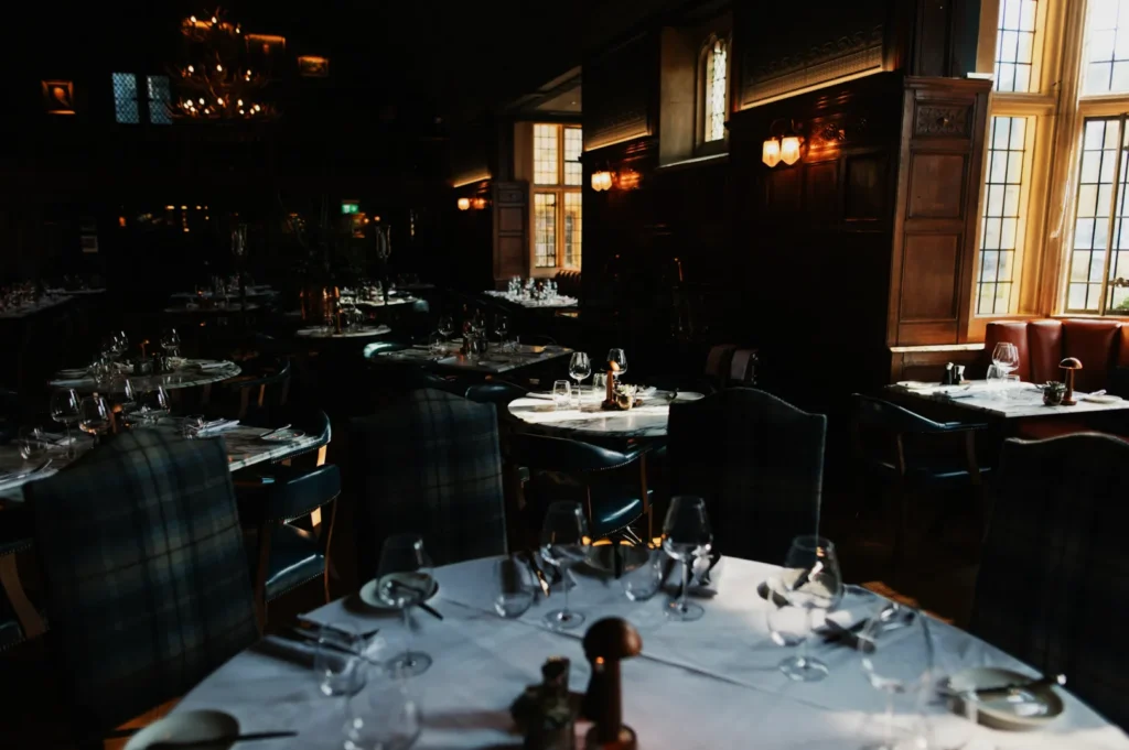 Dimly lit, elegant restaurant with round tables, white tablecloths, dark wood paneling, and a large chandelier, creating an upscale, cozy ambiance.