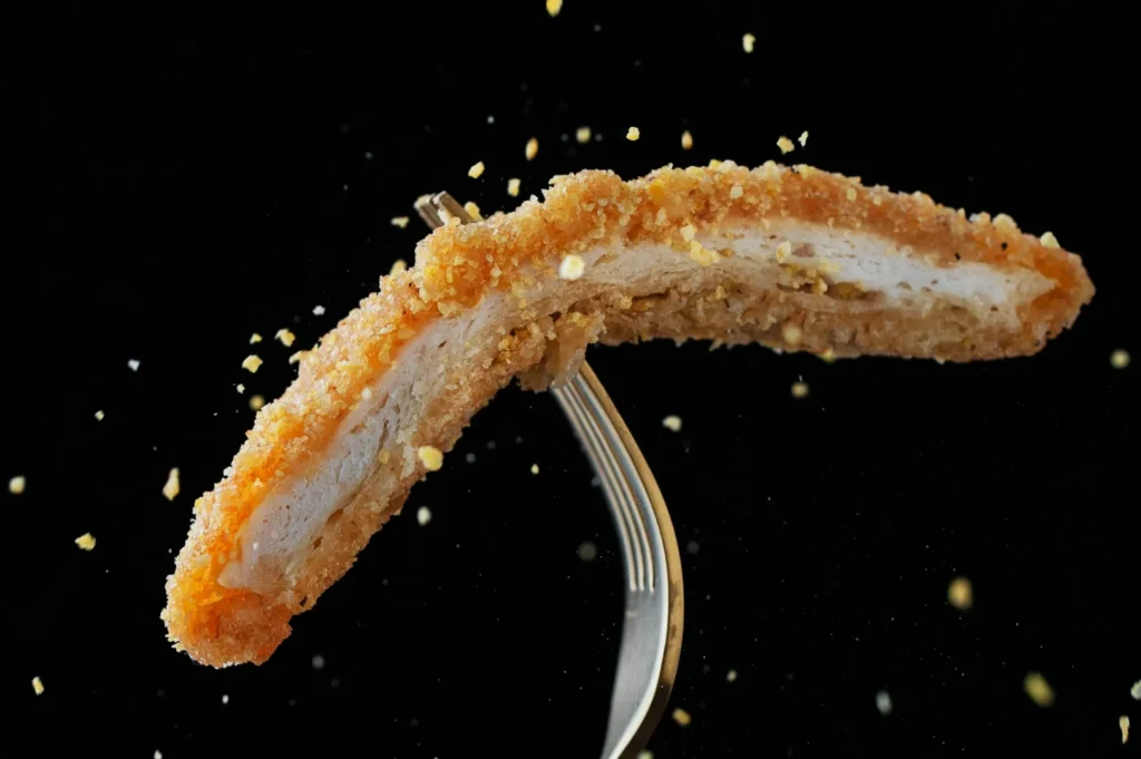 A crispy, breaded chicken strip is pierced by a fork, suspended in mid-air against a black background. Crumbs scatter dynamically around it.