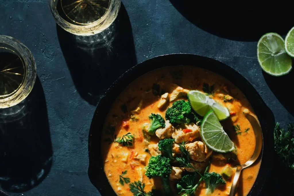 “A bowl of spicy curry with chicken, broccoli, cilantro, and lime slices on a dark table. Glasses of white wine and sliced lime are nearby.”