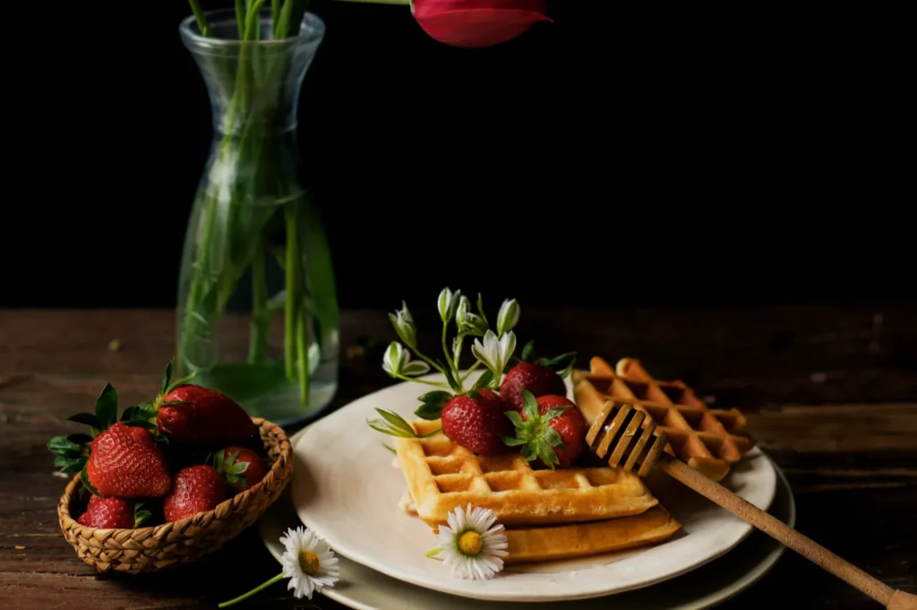 “A rustic still life with waffles topped with strawberries and small flowers on a white plate. A honey dipper rests on the plate. Nearby, a basket holds strawberries. A tall vase with a red tulip adds elegance.”