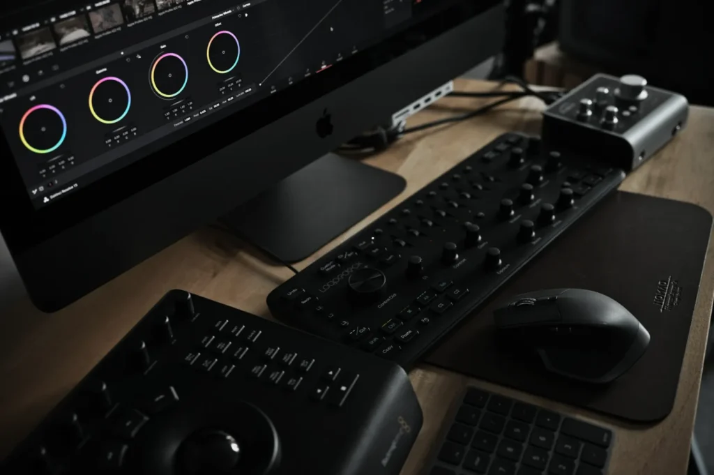 A editing workstation with a large monitor displaying color grading tools. A control panel, mouse, and audio interface are on a wooden desk.