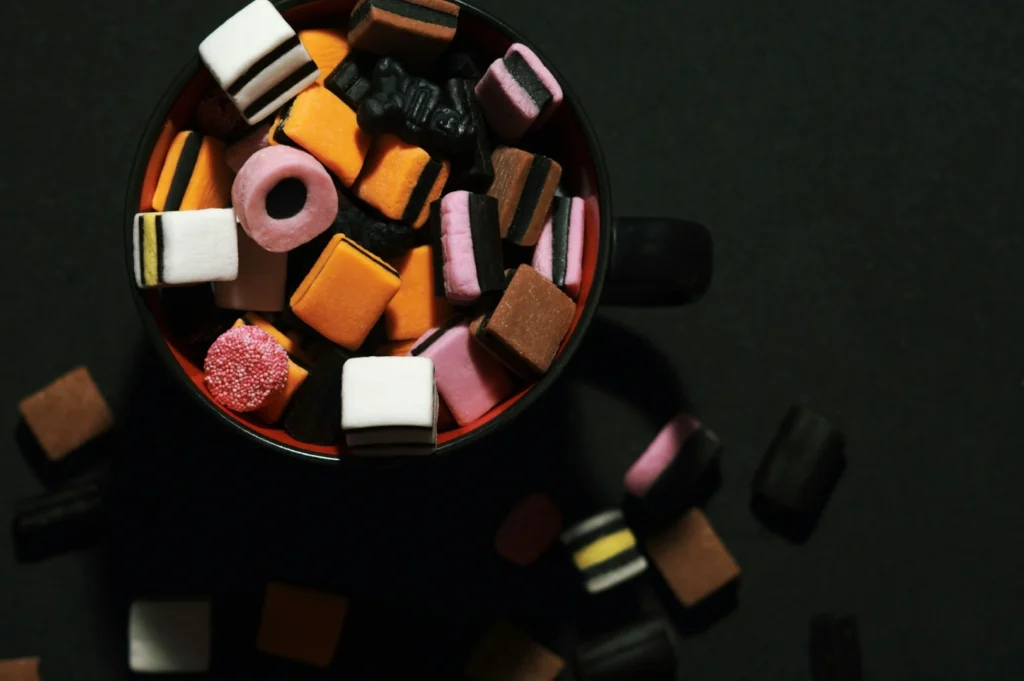 “A black cup filled with assorted, colorful licorice allsorts on a dark surface. Pieces are scattered outside, adding a playful, vibrant tone.”