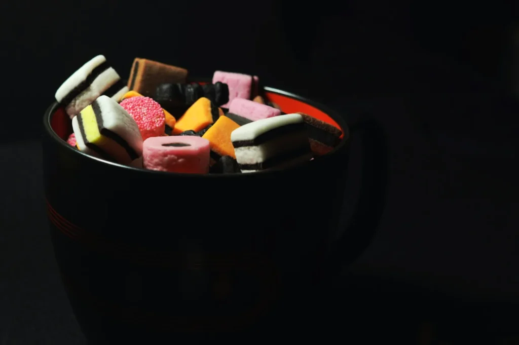 “A black mug filled with assorted colorful licorice allsorts, including pink, orange, black, and white pieces, on a dark background.”