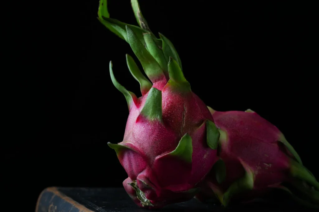 Two vibrant dragon fruits with pink skin and green spiky leaves sit against a dark background, highlighting their exotic texture and color.