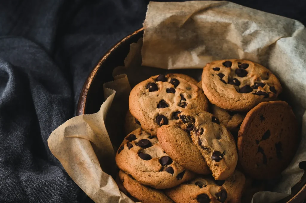 “A bowl of freshly baked chocolate chip cookies on crinkled parchment paper, set against a dark cloth background. The scene feels cozy and inviting.”