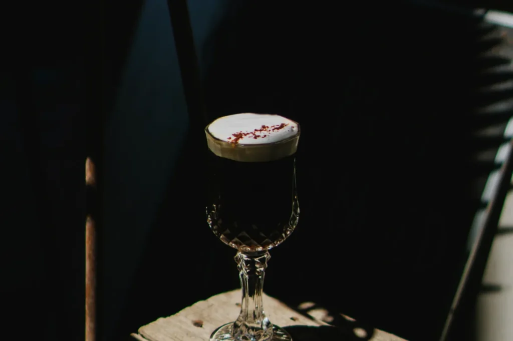 A vintage glass filled with dark Irish coffee topped with cream sits on a rustic wooden table, illuminated by soft, natural light creating a cozy ambiance.