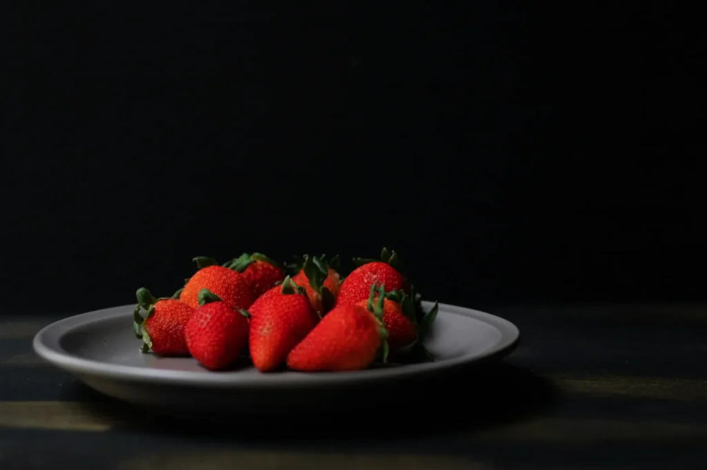 A gray plate with vibrant red strawberries sits on a dark surface against a black background. The scene conveys simplicity and freshness.