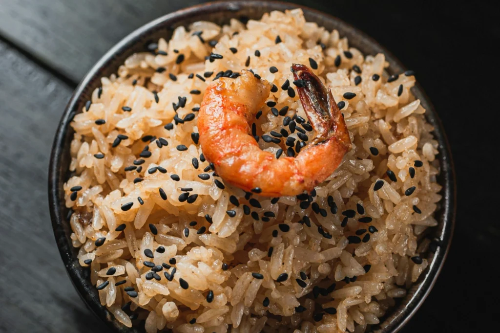 “A bowl of seasoned rice topped with a grilled shrimp and sprinkled with black sesame seeds, giving a savory and appetizing appearance.”