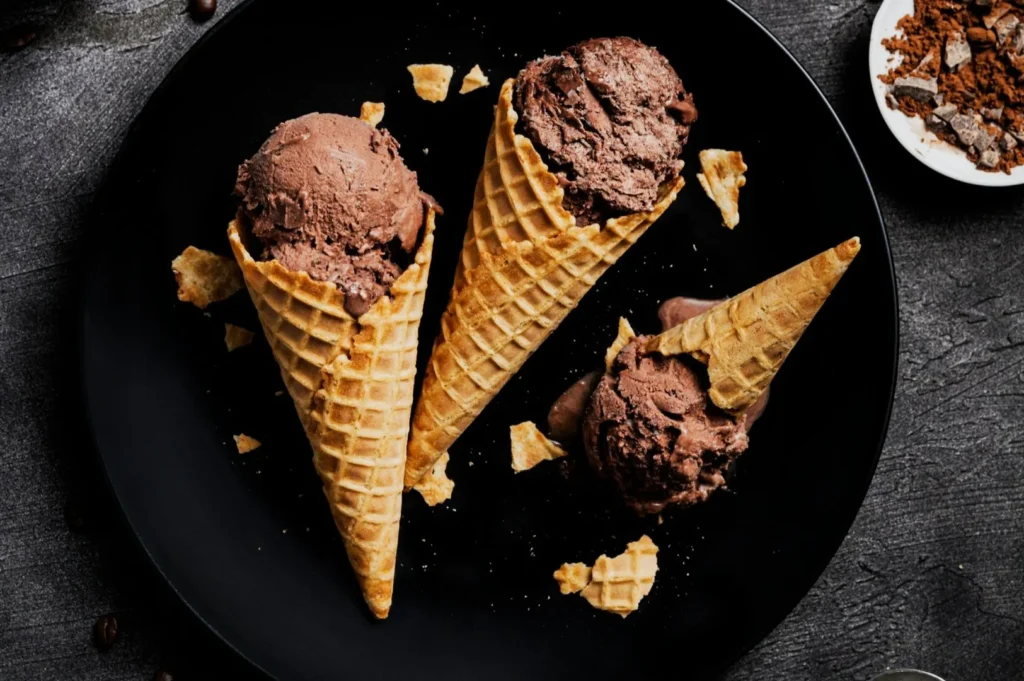 “Three waffle cones with scoops of chocolate ice cream on a black plate, surrounded by scattered waffle pieces, creating a tempting dessert scene.”