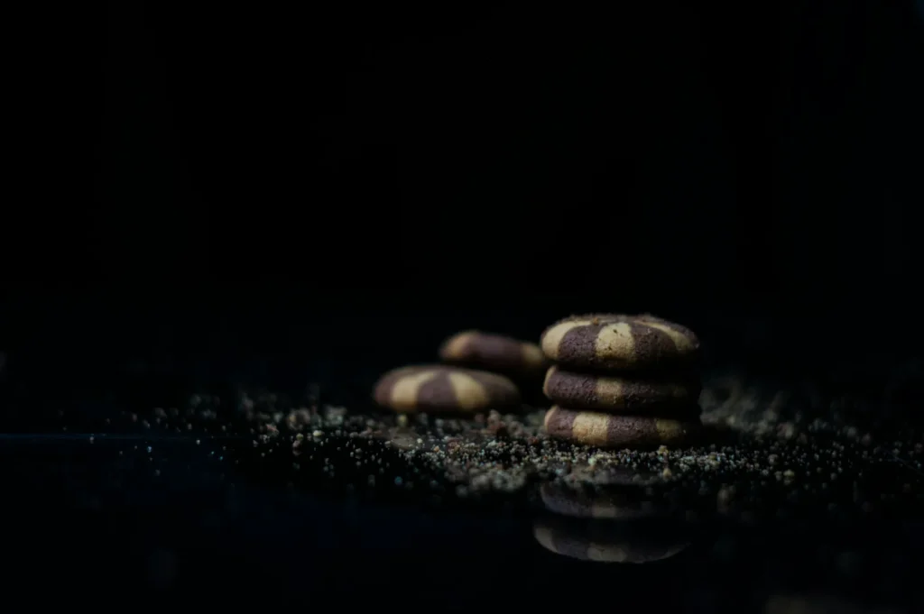 A stack of striped chocolate and vanilla cookies on a dark reflective surface, surrounded by scattered crumbs, against a black background, evokes a cozy, indulgent mood.