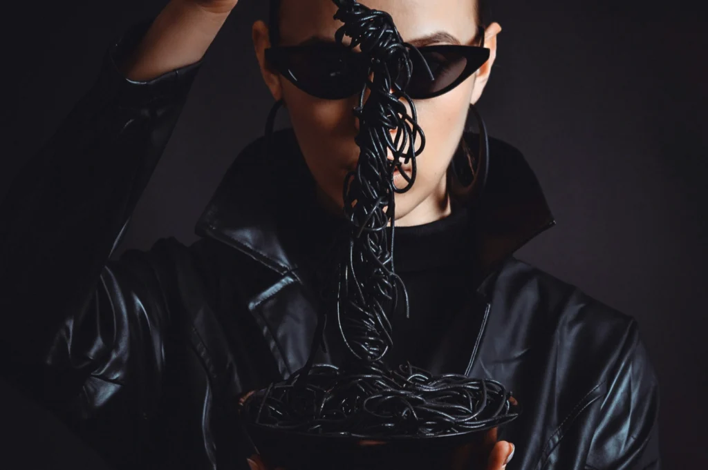 “A person in a black leather jacket and sunglasses holds a fork with black noodles above a bowl. The dramatic, stylish scene conveys a mysterious vibe.”