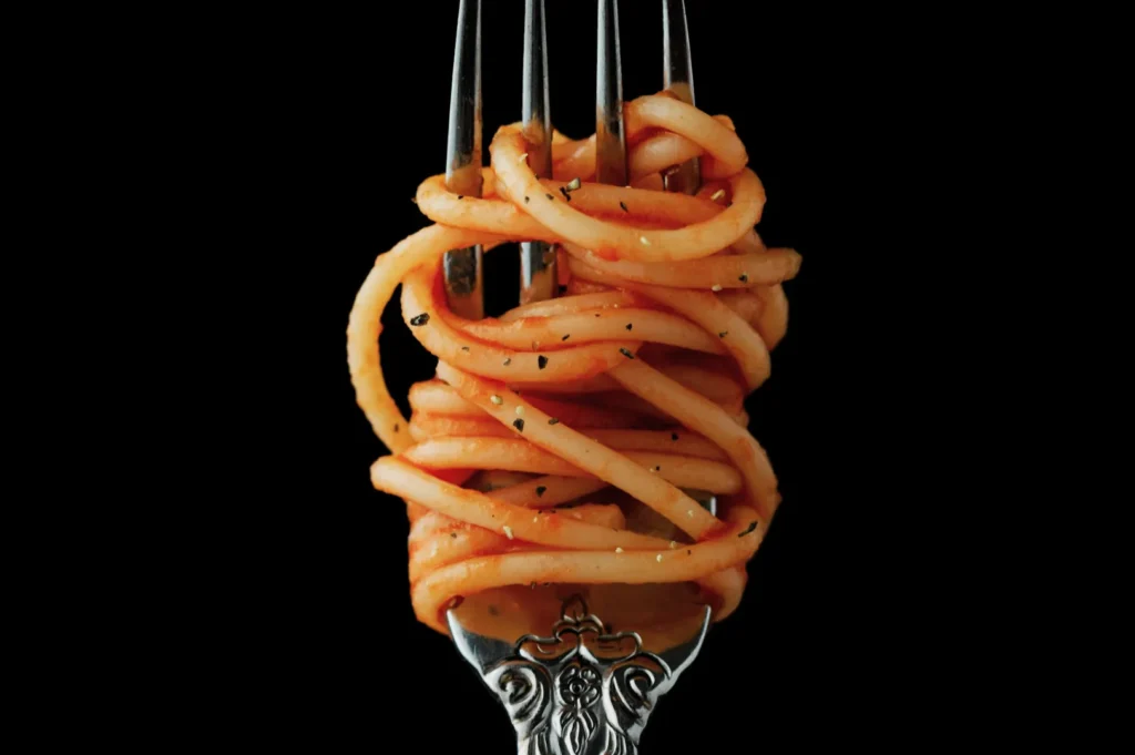 “A fork with ornate detailing holds neatly twirled spaghetti coated in red sauce, sprinkled with herbs, set against a black background.”