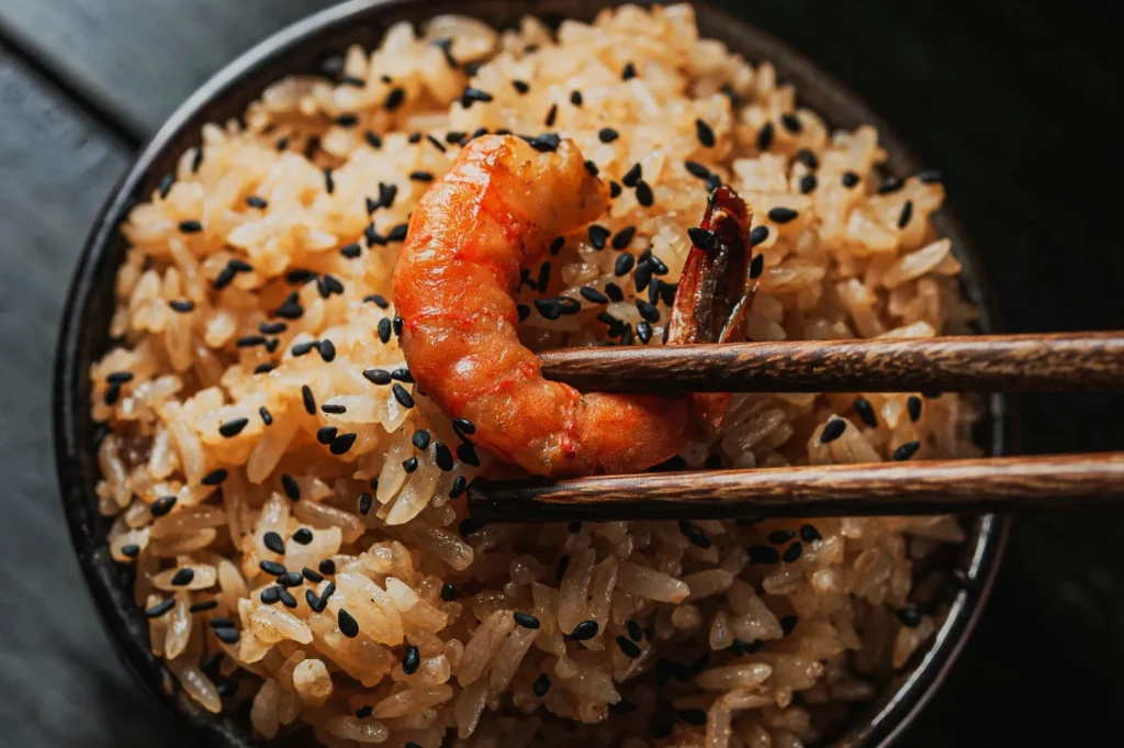 “Close-up of a bowl of rice topped with black sesame seeds, and a grilled shrimp being held by chopsticks. The scene conveys appetizing warmth.”