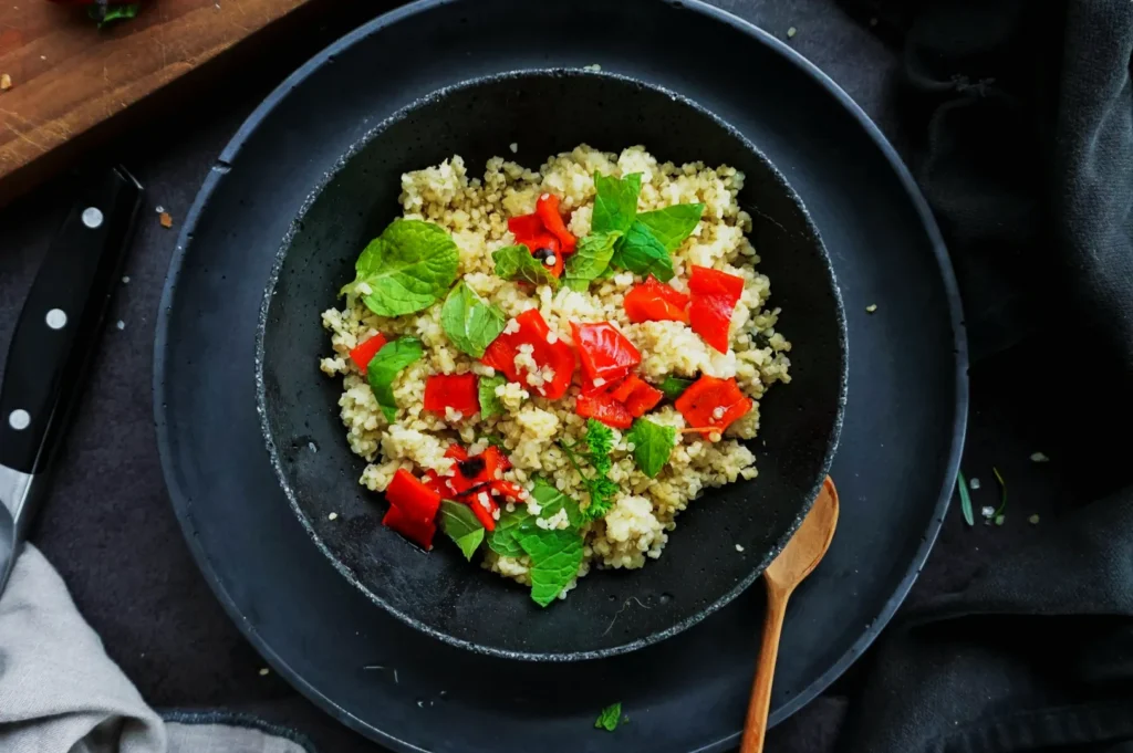 Quinoa salad on a black plate, garnished with vibrant red bell peppers and fresh green mint leaves, next to a wooden spoon and knife.