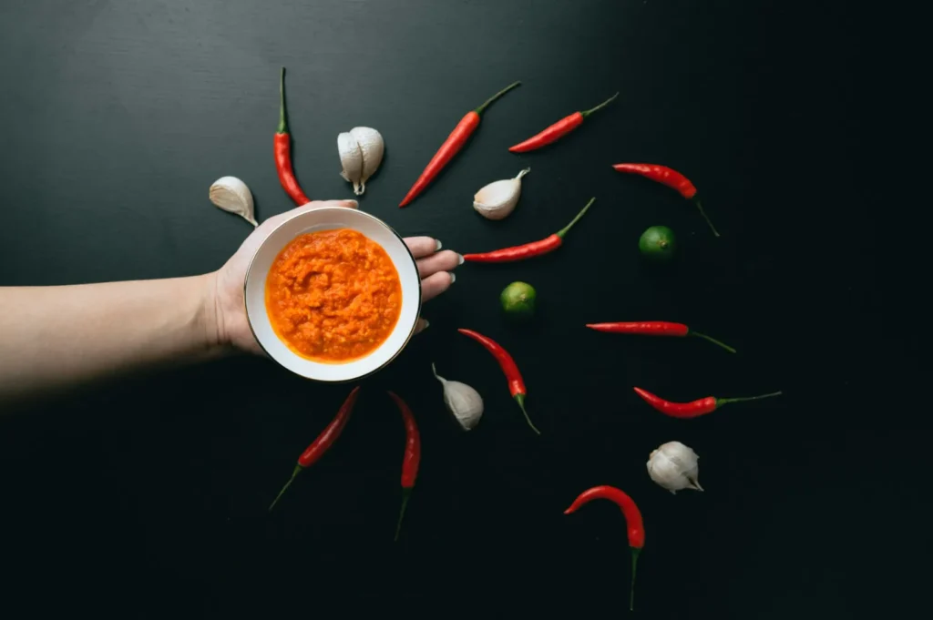 “A hand holds a bowl of red chili sauce against a black background. Surrounding ingredients include red chilies, garlic cloves, and small green limes, creating a vibrant and spicy theme.”
