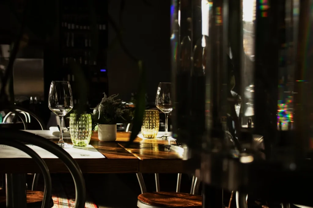 A dimly lit restaurant table with wine glasses and green textured drinking glasses. Sunlight casts soft shadows, creating an intimate, warm atmosphere.